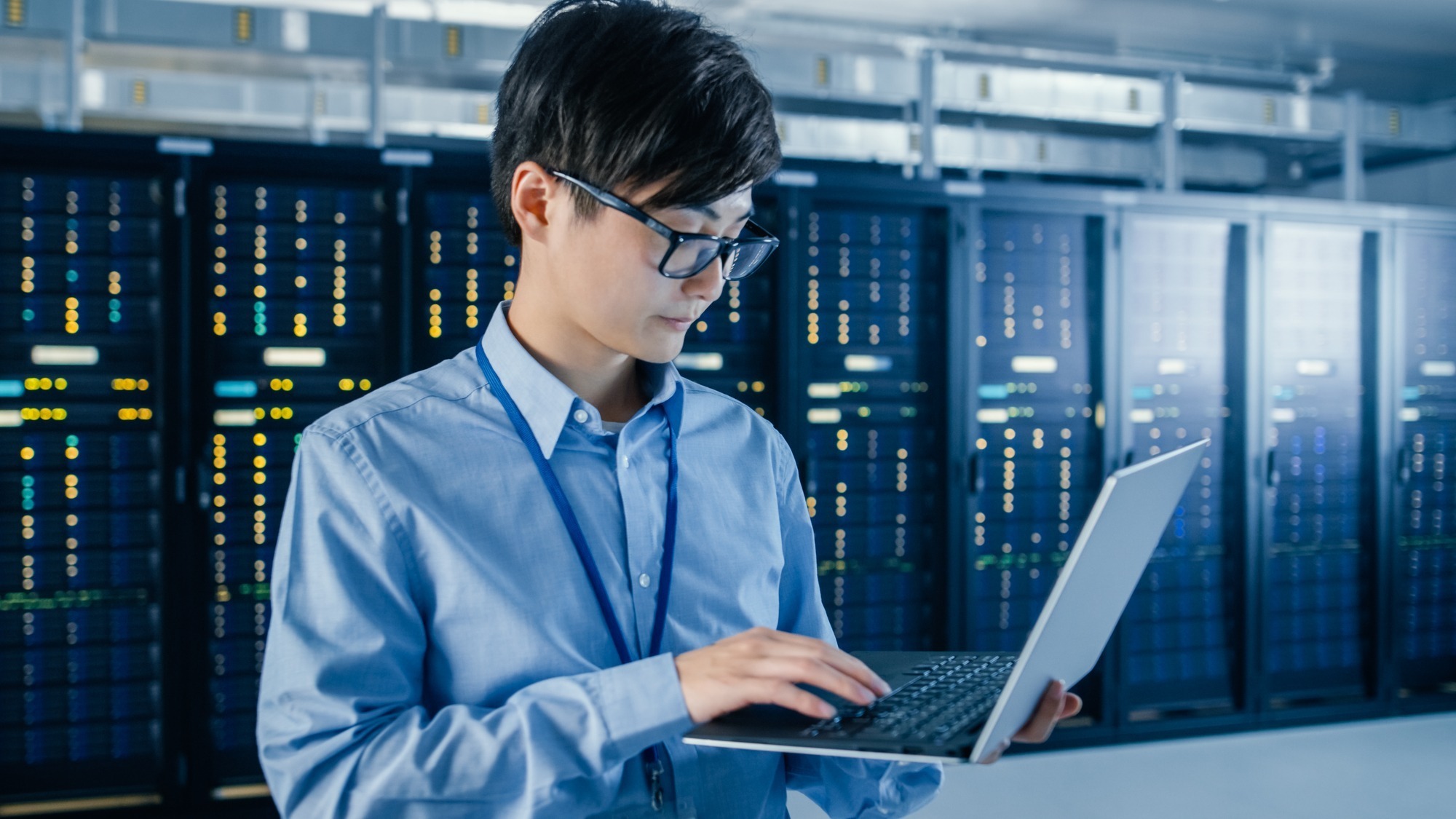 In the Modern Data Center: Portrait of IT Engineer Standing with man in glasses in server room working on a laptop at IT service company