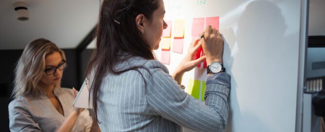 Waterfall project management example showing two professionals organizing tasks on a whiteboard with color-coded sticky notes arranged in sequential phases.