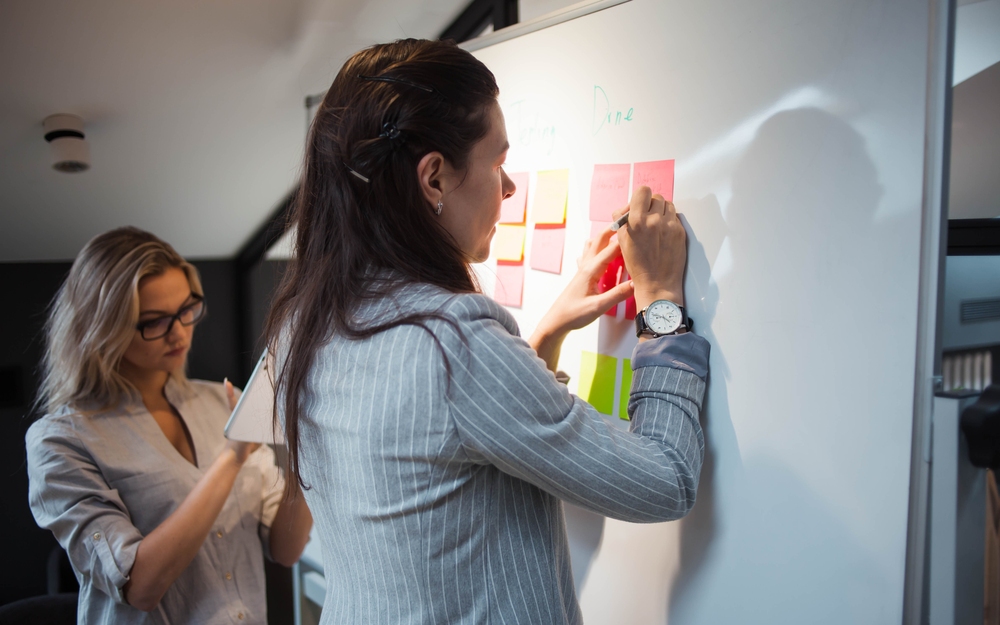 Waterfall project management example showing two professionals organizing tasks on a whiteboard with color-coded sticky notes arranged in sequential phases.